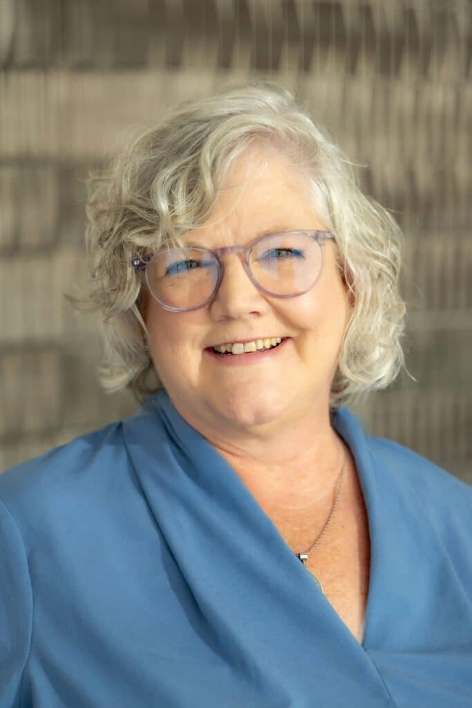 Professional headshot of a smiling woman with short curly gray hair and glasses, wearing a light blue blouse, in front of a softly blurred neutral background.